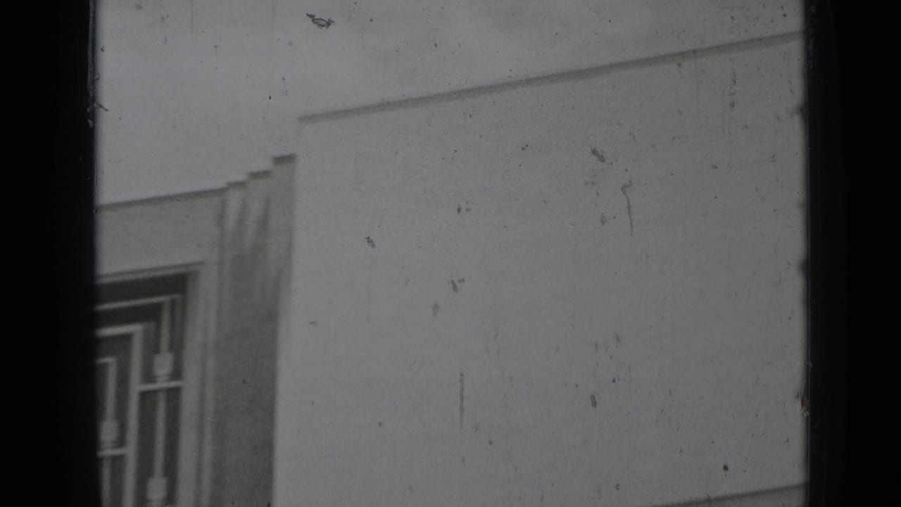 Black-and-white 8mm frame showing part of a geometric window, pale wall surface, and pronounced archival film texture.