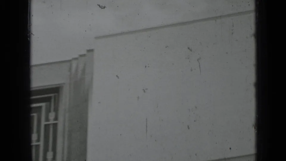 Black-and-white 8mm frame showing part of a geometric window, pale wall surface, and pronounced archival film texture.