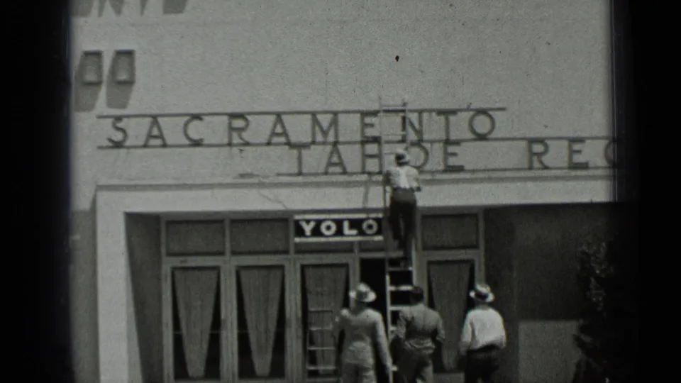 1937 San Francisco 8mm frame with men in hats gathered on the sidewalk while a ladder worker handles signage above storefront doors.