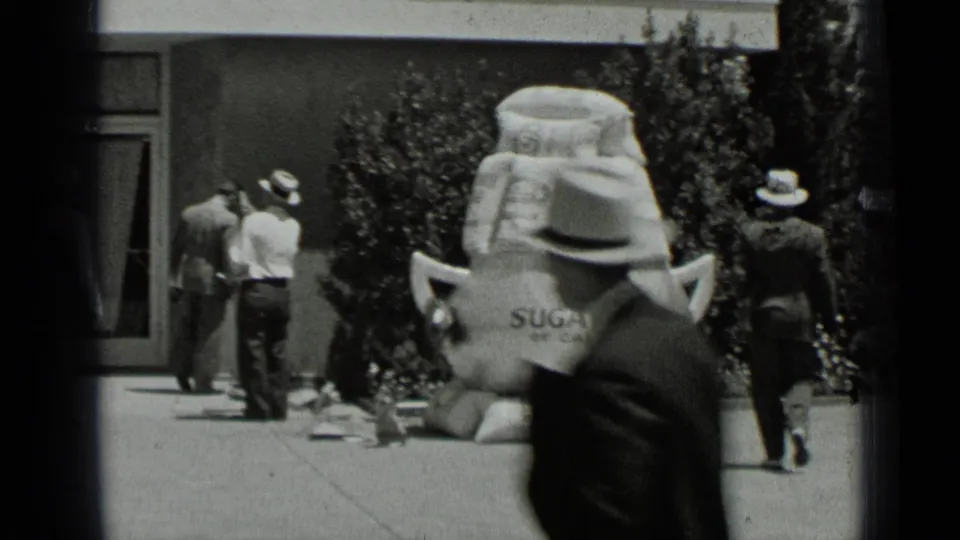 1937 San Francisco frame showing men near the storefront facade, a ladder worker, and a large promotional sugar-bag display on the sidewalk.