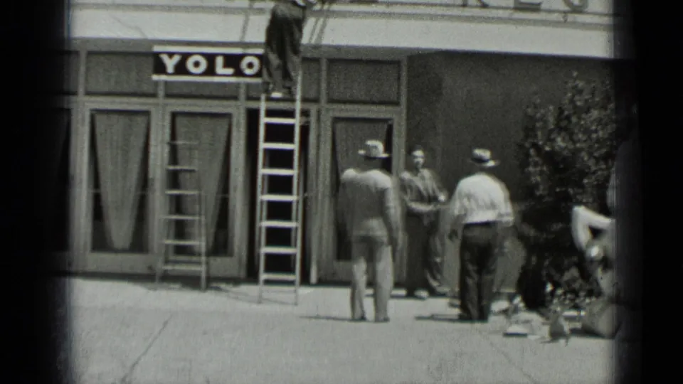 1937 San Francisco storefront scene with a man on a ladder working near a YOLO sign as men watch from the sidewalk.