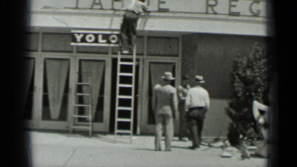 Men in hats stand near ladders and sidewalk materials outside a San Francisco storefront with YOLO lettering in 1937 footage.