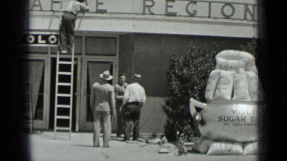 1937 San Francisco frame showing men, a ladder, partial YOLO signage, and the edge of a large sugar-bag display outside a storefront.