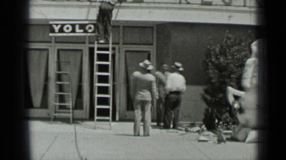 1937 San Francisco street scene with a man on a ladder adjusting a sign above a YOLO storefront as suited men observe.