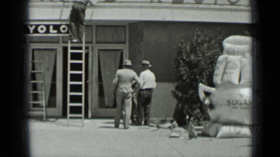 Two men stand beneath the YOLO sign and ladder, with storefront doors and a large sugar sack display visible at the edge.