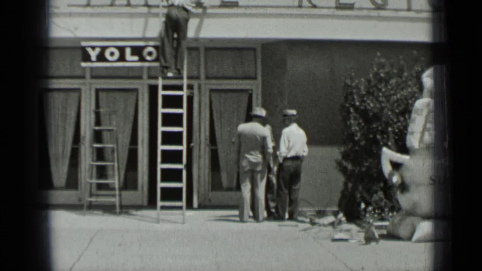 In the final frame, two men face the storefront beneath the YOLO sign while the ladder and sidewalk materials remain visible.
