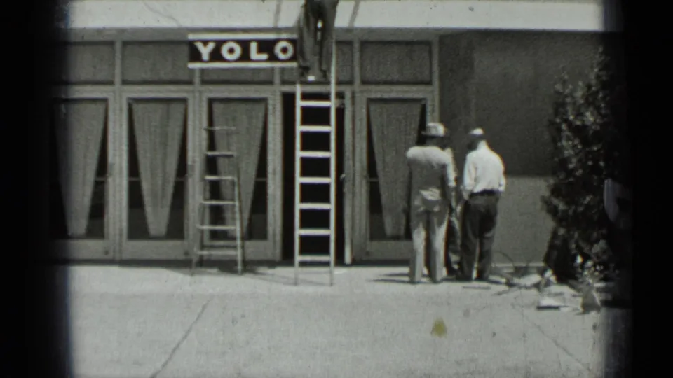 1937 San Francisco 8mm frame showing men in hats near ladders outside a Yolo storefront, with the camera looking across a concrete sidewalk.