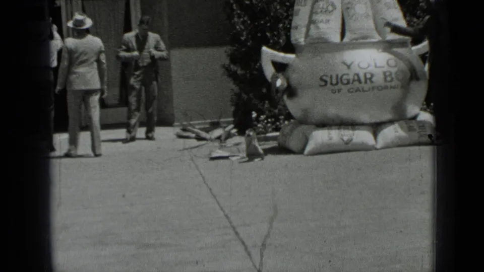 Black-and-white 8mm frame from 1937 San Francisco showing men on a sidewalk beside the Yolo Sugar Bowl display and storefront area.