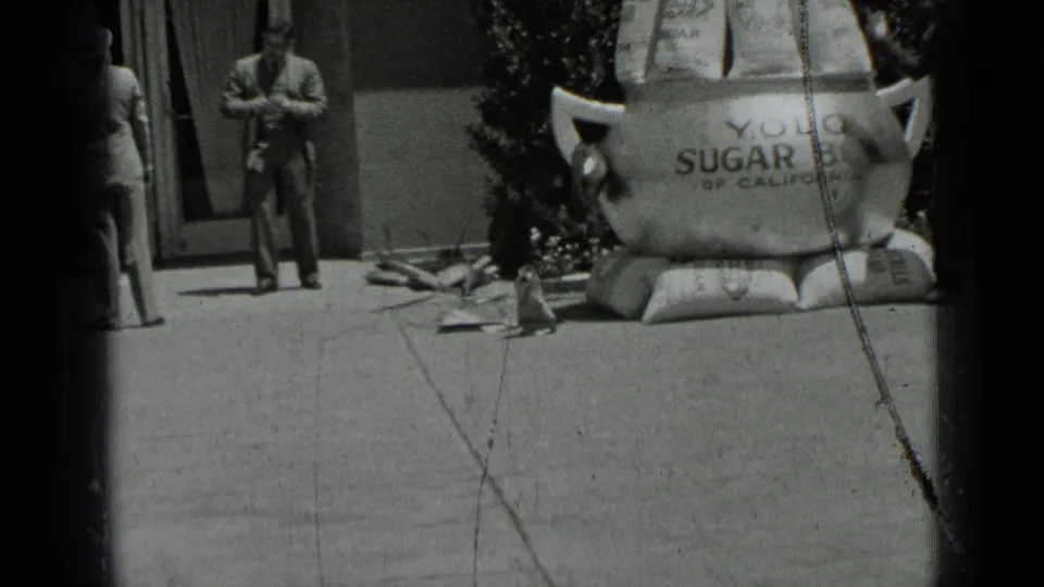1937 San Francisco frame with a suited man walking near the Yolo Sugar Bowl display, shrubs, sacks, and visible film scratches.