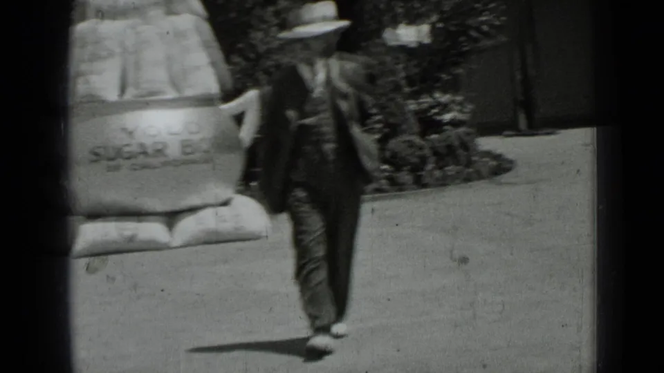 A blurred 1937 San Francisco frame shows a man in a hat walking past the oversized sugar bowl prop and shrub-lined sidewalk.