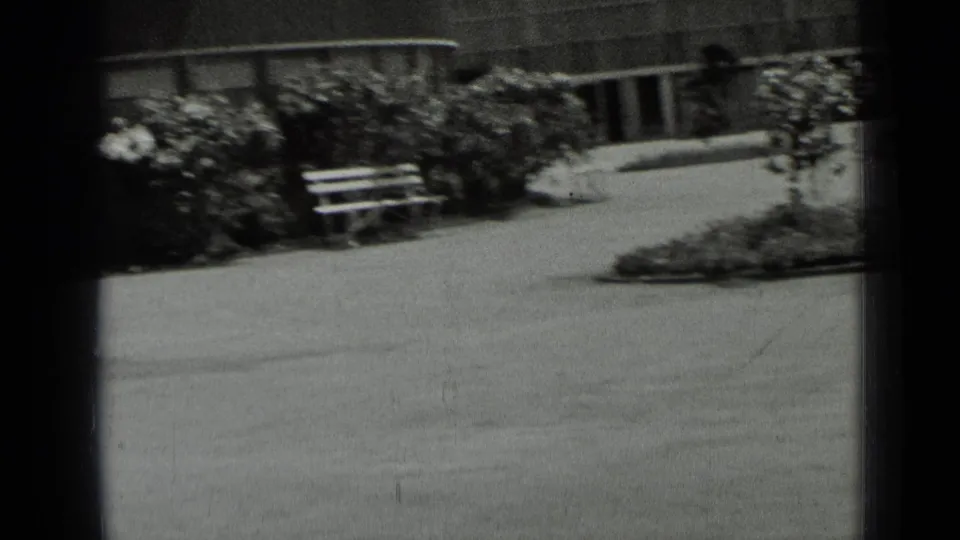 1937 San Francisco home-movie frame showing a paved plaza area, benches, shrubs, and the edge of a public-looking building.