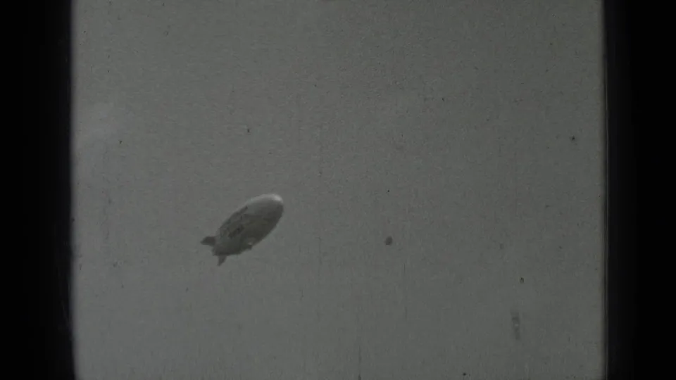The blimp remains small against a gray sky in archival 8mm footage, with dust marks, scratches, and dark frame borders.