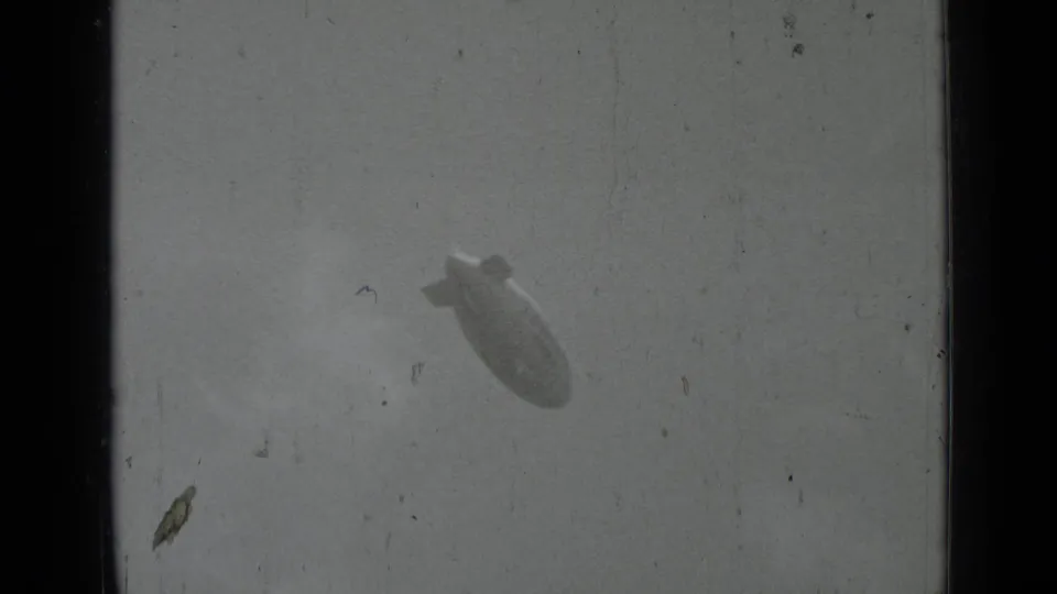 Distant Goodyear blimp appears angled in the sky within a worn 8mm film frame.