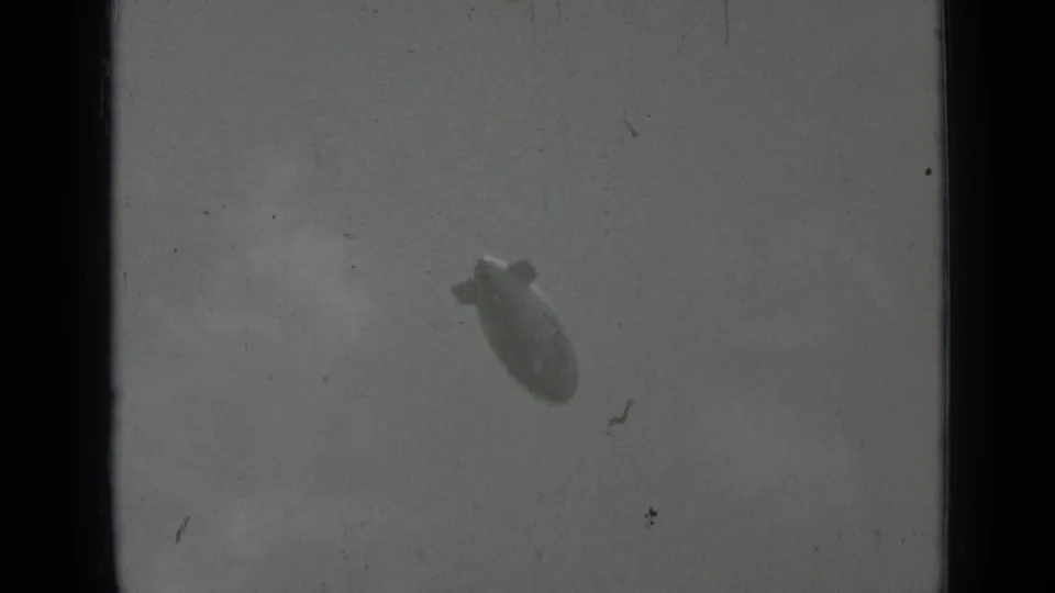 Soft 8mm frame showing the advertising blimp drifting through a hazy sky with visible film wear.
