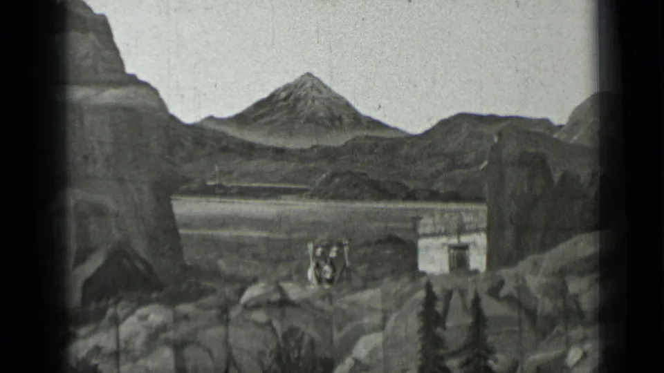 1937 8mm frame showing a rocky mountain backdrop, small building, and a group of distant performers in a staged outdoor scene.