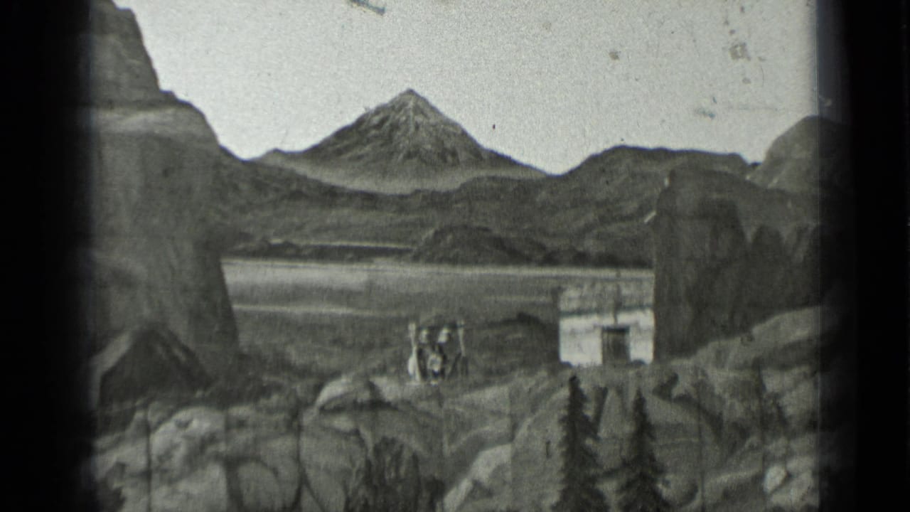 Rocky landscape with distant people beside a small building and mountain backdrop.