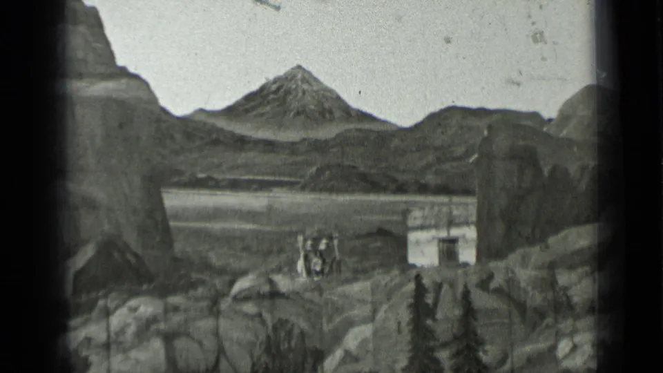 1937 home-movie frame with distant figures, a small rectangular set piece, and a prominent mountain backdrop within the 8mm gate.