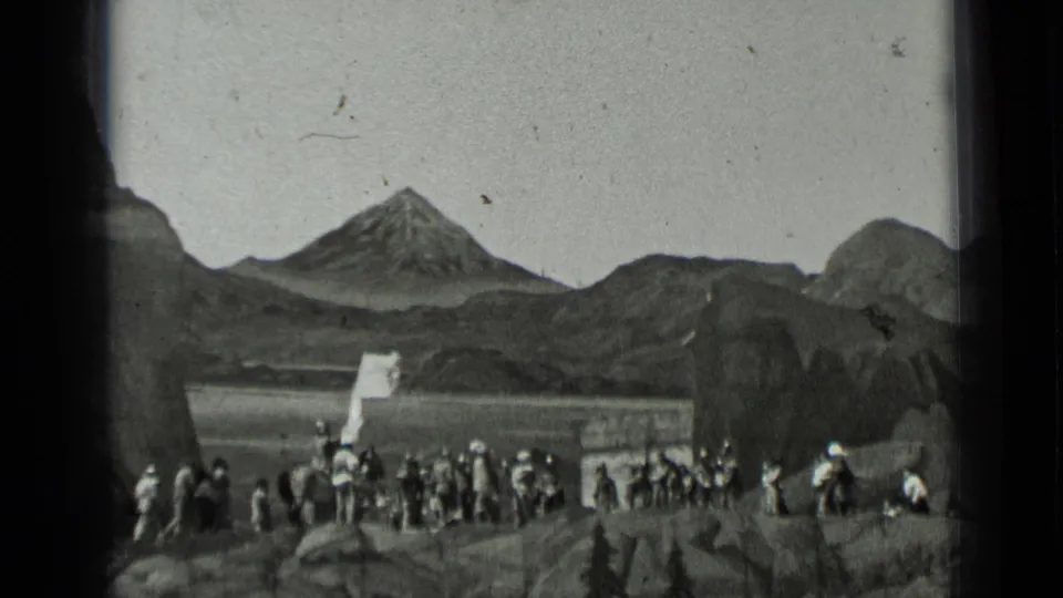 1937 8mm frame showing men in dark uniforms clustered near foreground rocks and a mountain-shaped backdrop.