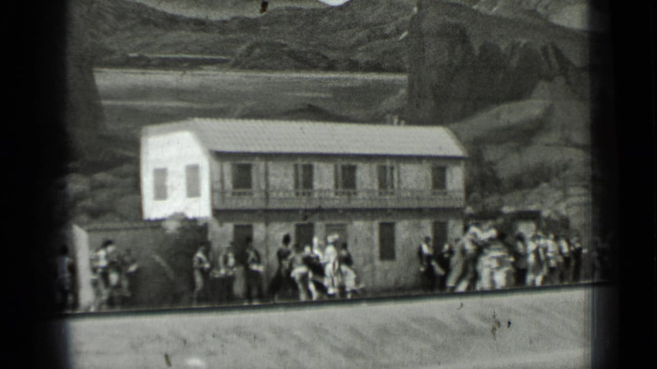 Crowd gathered and dancing outside a two-story building with painted mountains behind it.