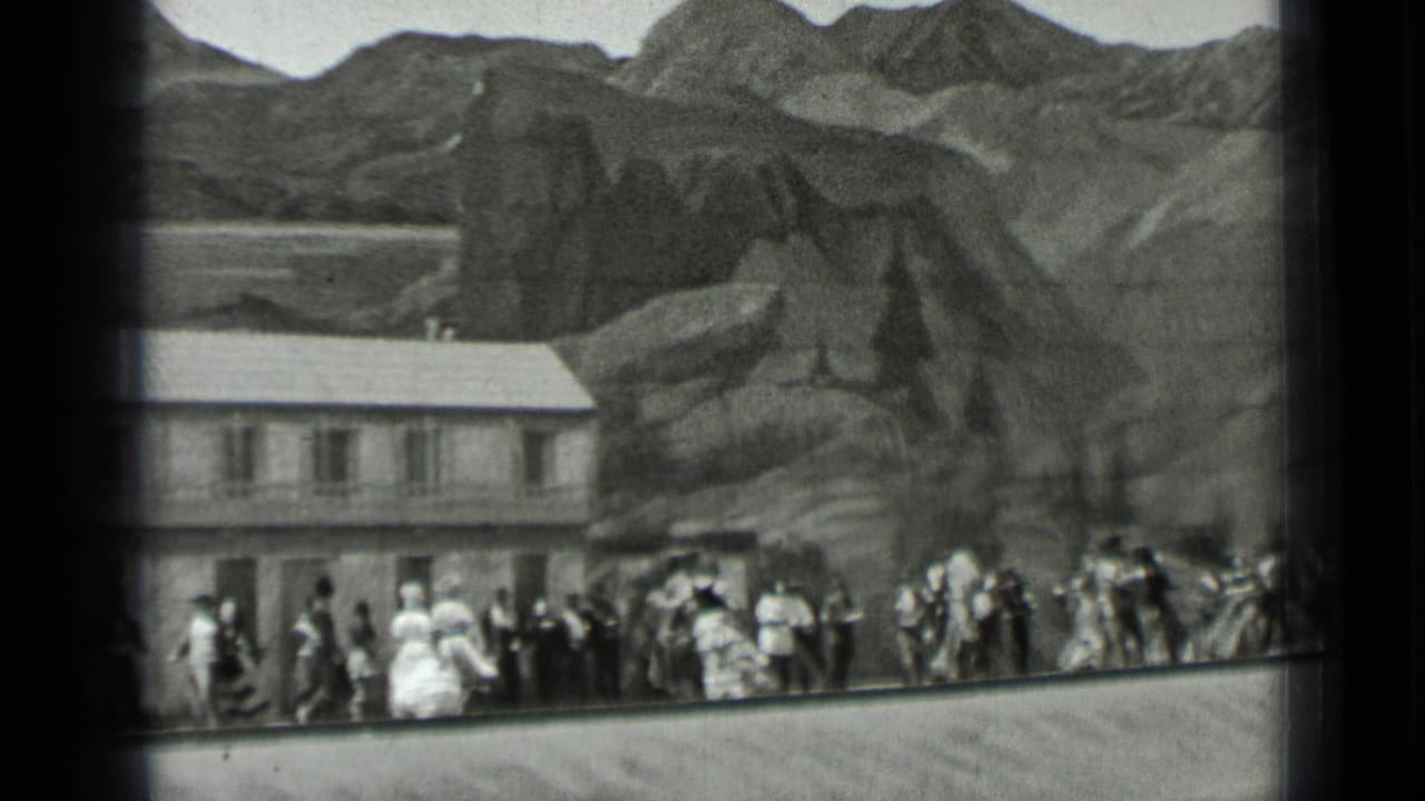 Line of people dancing and watching in front of a long building and rugged backdrop.