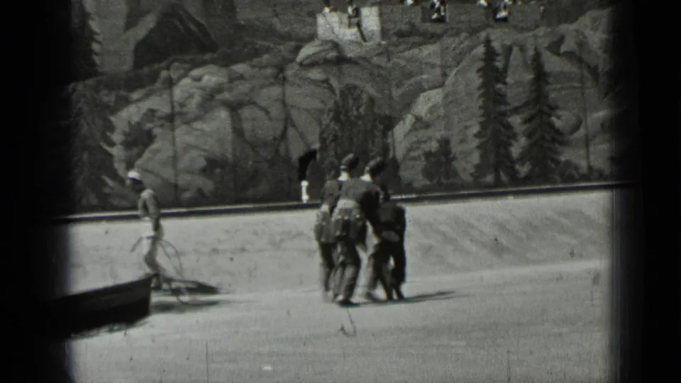 In 1937 San Francisco home-movie footage, dark-uniformed figures walk past a painted rocky forest backdrop while another figure stands at left.
