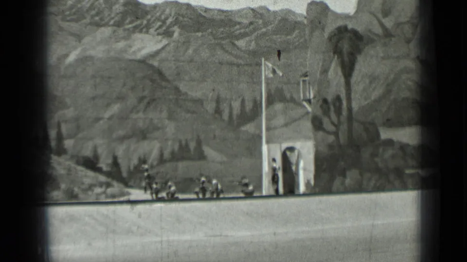 1937 San Francisco 8mm frame showing uniformed figures and a flag near a small building against a painted rocky mountain backdrop.