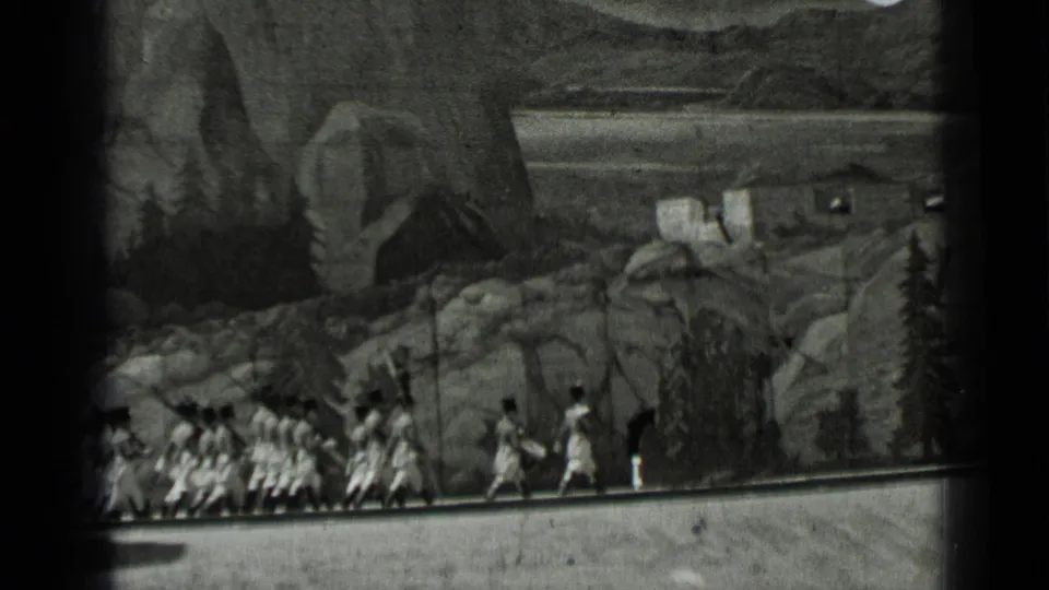1937 San Francisco 8mm frame showing a line of uniformed marchers moving past rocks, trees and set buildings in a staged landscape.