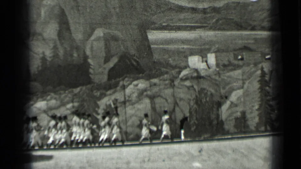 1937 San Francisco 8mm frame of uniformed figures marching through a staged rocky mountain landscape with small buildings and trees.