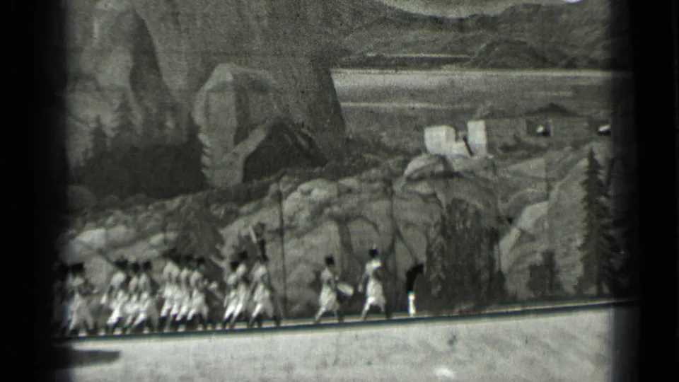Grainy 8mm view of period-costumed extras moving across a backlot-style set with painted landscape scenery in the background.