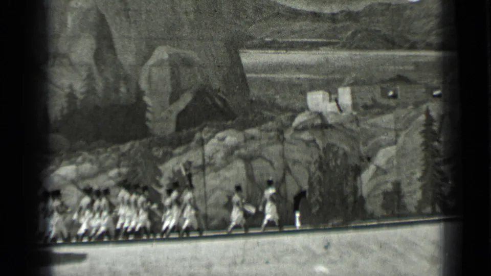 Final frame shows extras on a studio walkway before painted rocky scenery and small cabin-like forms, with visible 8mm frame edges.