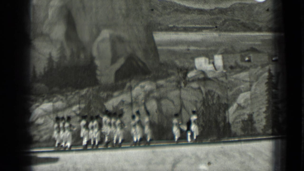 A larger formation of uniformed marchers crosses the stage from left to right.