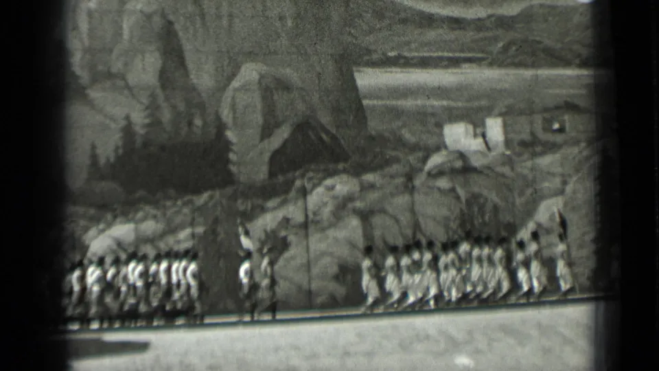 1937 San Francisco home-movie frame showing uniformed marchers and a dark-clad leader or conductor before a painted mountain set.