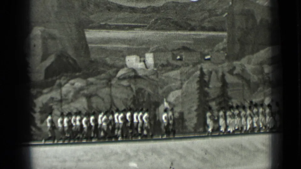 Distant 8mm view of uniformed marchers moving in formation before rocky scenery, with a leader or standing figure near the line.