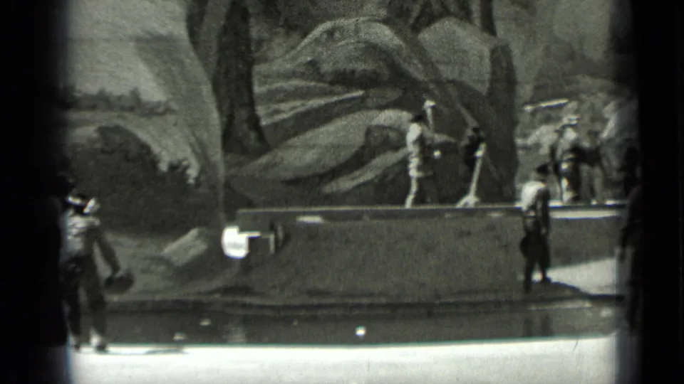 1937 San Francisco home-movie frame showing a standing worker, background riders, and laborers near rocky terrain and a paved or wet edge.