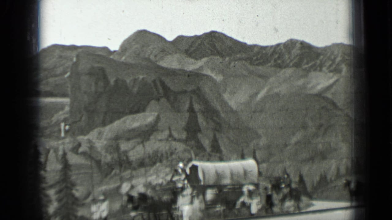 Covered wagon caravan with people and horses below rugged mountains.
