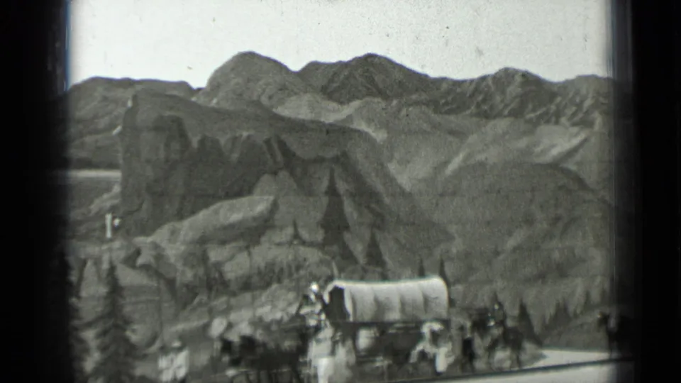 1937 8mm frame showing covered wagons, horses, and small groups of people moving through a mountainous western-style scene.