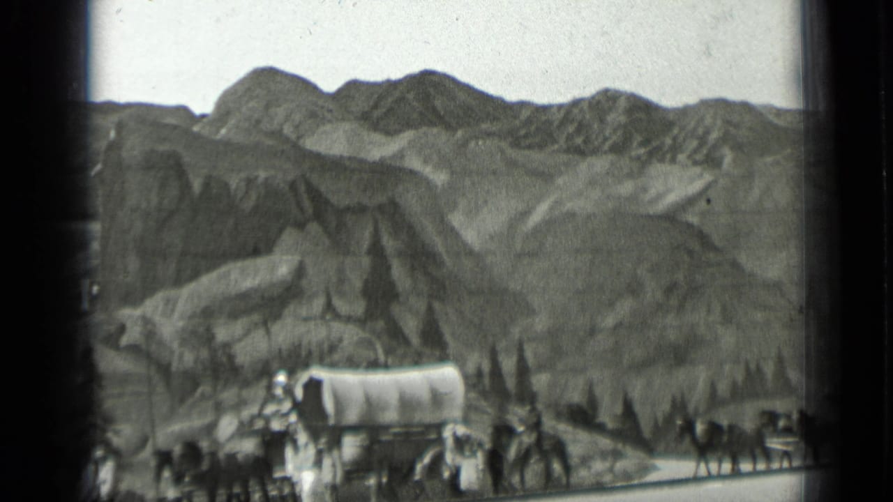 Horse-drawn covered wagon and riders in front of mountain scenery.