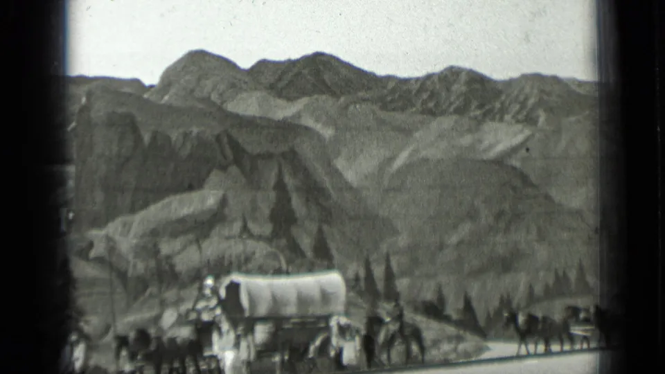 Black-and-white 8mm frame with a covered wagon, horses, and people traveling across a rugged mountainous backdrop.
