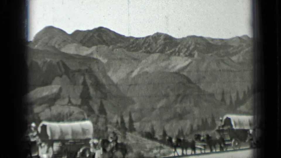 1937 home-movie frame showing wagons, horses, and people progressing through a western frontier-style mountain setting.