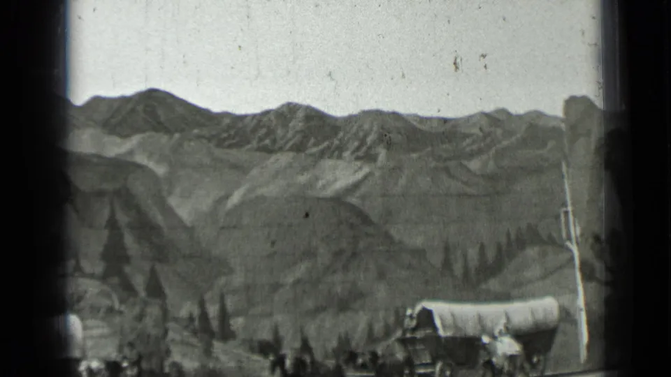 Black-and-white 8mm frame of a covered wagon group and horses near a path, with ridges and sparse trees in the background.