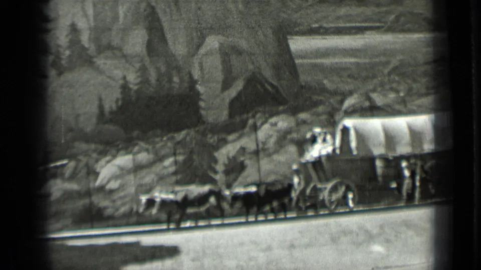Black-and-white 8mm frame of a covered wagon and animals crossing a diorama-like western scene with rocks, trees, and painted scenery.