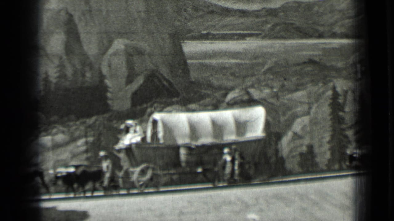 Covered wagon with oxen and small traveler figures before a painted mountain backdrop.