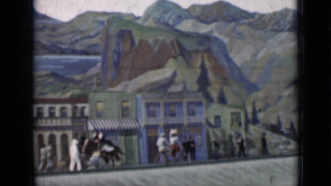1937 San Francisco 8mm frame showing small figures on a sidewalk before painted storefronts and a mountain backdrop.