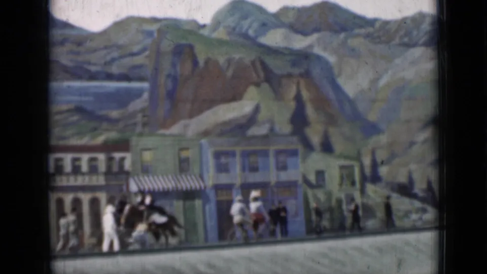 1937 San Francisco 8mm frame showing small figures on a sidewalk before painted storefronts and a mountain backdrop.