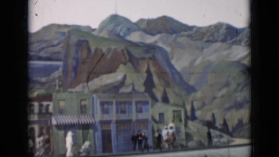 1937 San Francisco home-movie frame with figures gathered near storefront facades and a dramatic painted mountain background.