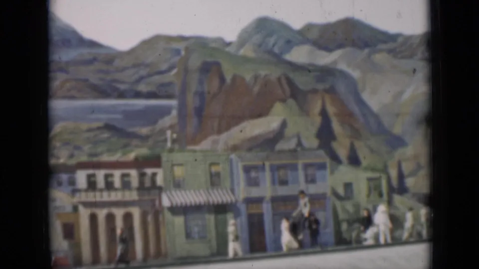 1937 San Francisco 8mm frame showing figures near a painted streetscape with hills, water, and mountain scenery behind.