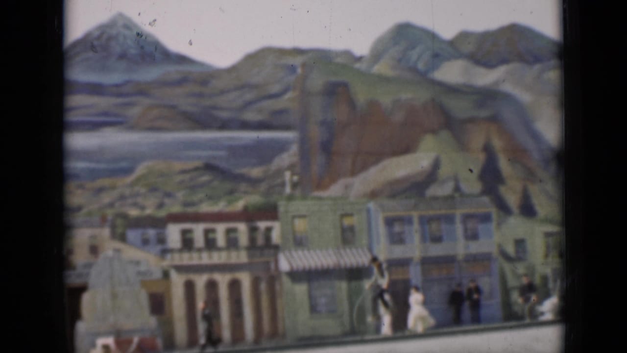 Painted storefronts and mountain backdrop with people near high-wheel bicycles.