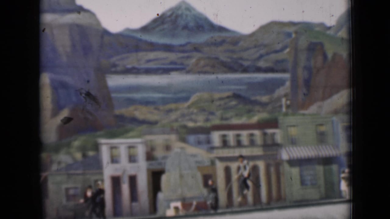 A rider on an old-fashioned high-wheel bicycle moves through a staged San Francisco streetscape with painted scenery behind.