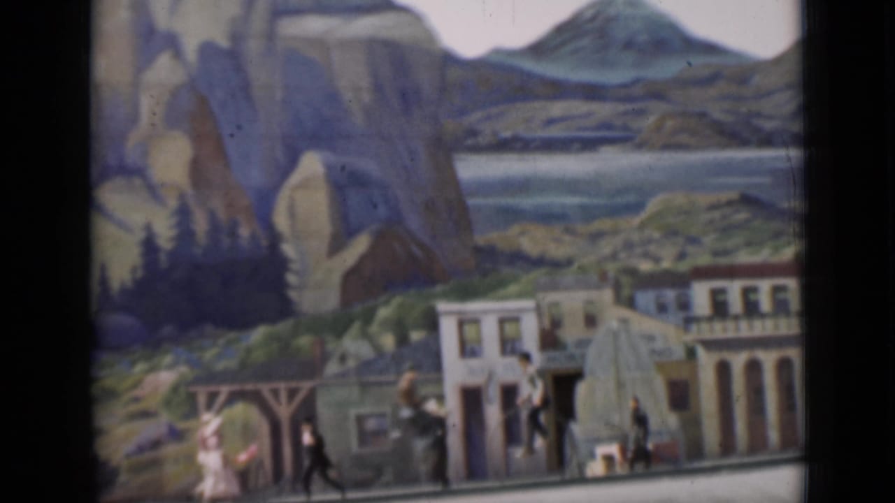 1937 home-movie frame with high-wheel bicycle riders and pedestrians passing a staged street set before painted rocks and hills.