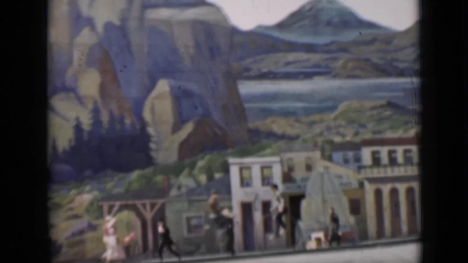 1937 home-movie frame with high-wheel bicycle riders and pedestrians passing a staged street set before painted rocks and hills.